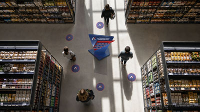 Overhead view of shoppers walking between tall grocery shelves, with a blue directional sign and circular A-floor markers.