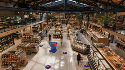 A large modern supermarket seen from above, with tall shelves, two balcony levels, and shoppers browsing the aisles.