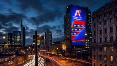Night city skyline at dusk with a busy road in the foreground, car light trails, and a tall building displaying a bright digital billboard.